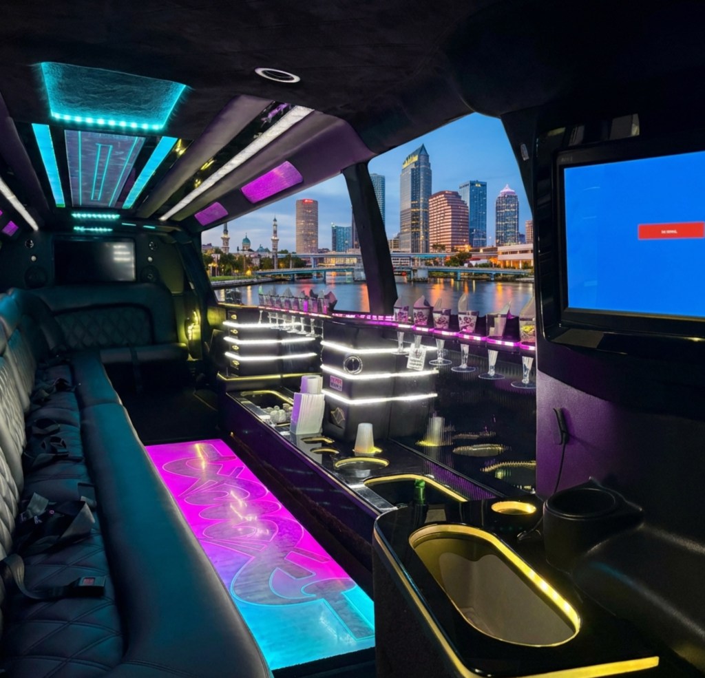 Interior of a luxury limousine featuring plush seating, modern lighting, a bar area with glasses, and a view of a city skyline at dusk.