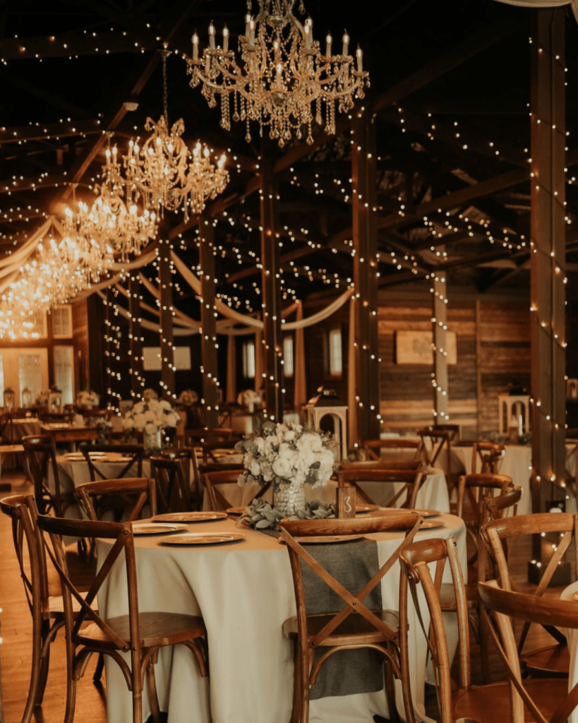 Elegant dining area with wooden chairs, adorned tables, and decorative floral centerpieces, illuminated by hanging chandeliers and string lights.
