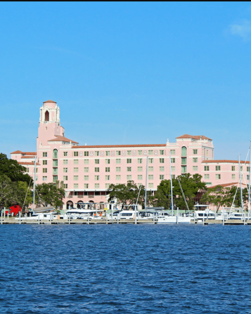 View of a large pink hotel building by the waterfront, featuring a clock tower and surrounded by boats docked at a marina, under a clear blue sky.