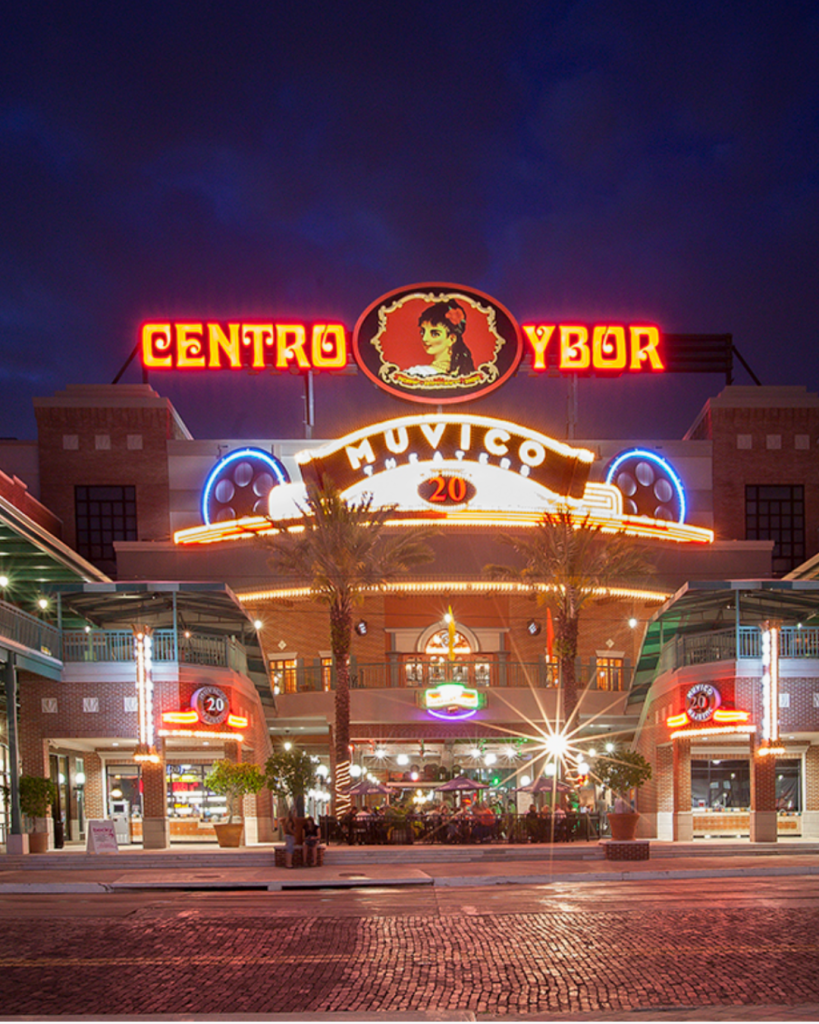 Exterior view of Centro Ybor with neon lights at night, featuring Muvico Theater signage and palm trees.