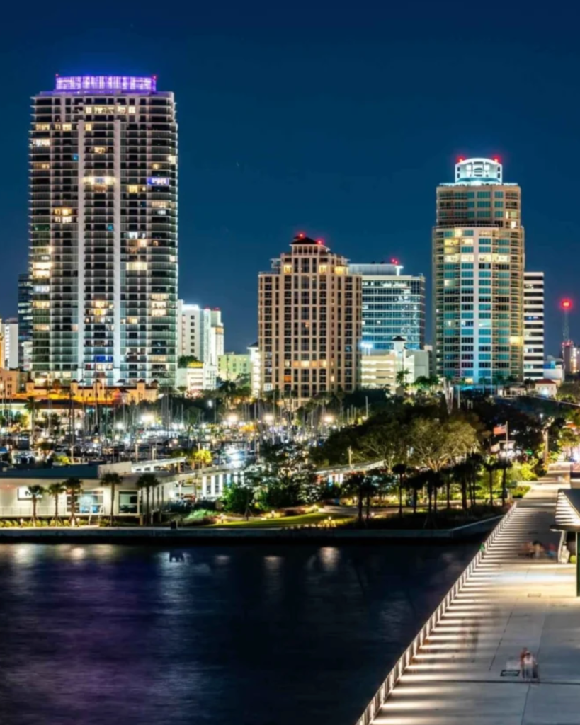 City skyline at night featuring illuminated skyscrapers and a waterfront promenade.
